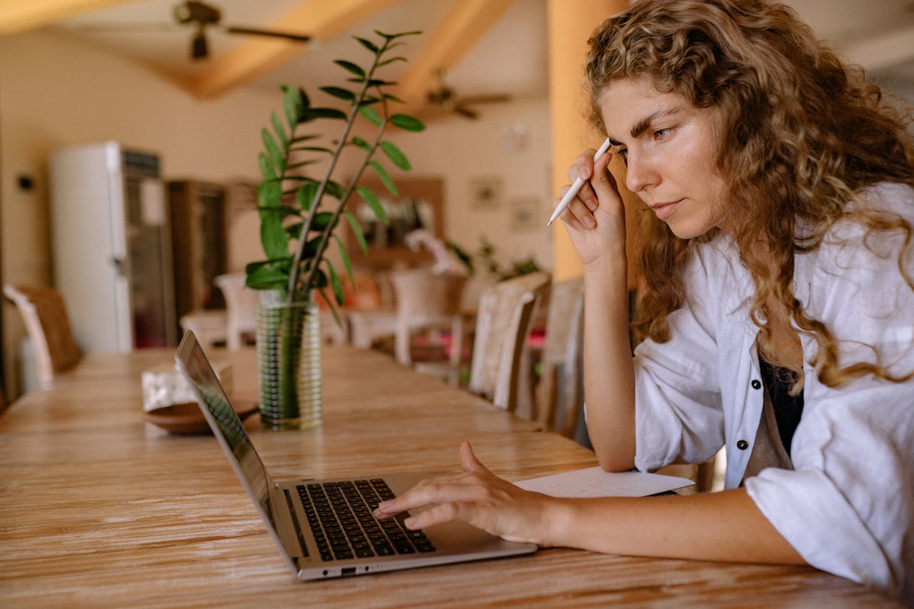 A woman working at a table with her laptop while holding a pen at her head, focusing on web design and digital tasks, representing productivity.