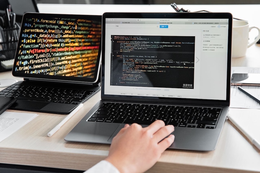 Developer writing code on a laptop, with lines of programming code displayed on both a laptop and a tablet screen on a office desk.